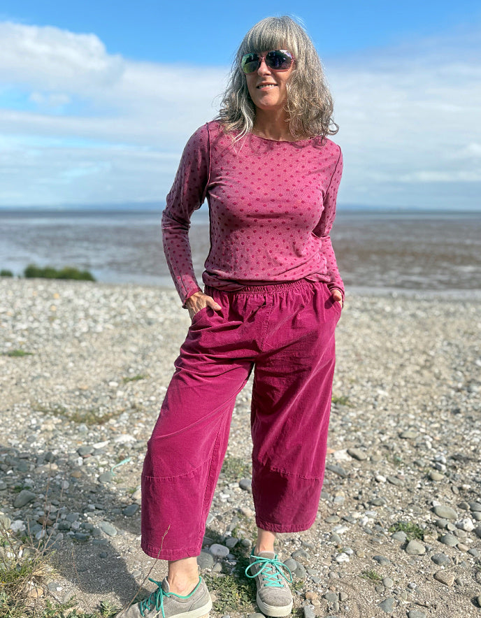 Woman in pink needlecord lantern trousers and a pink pink spotty top standing on a pebbly beach with ocean and sky in the background