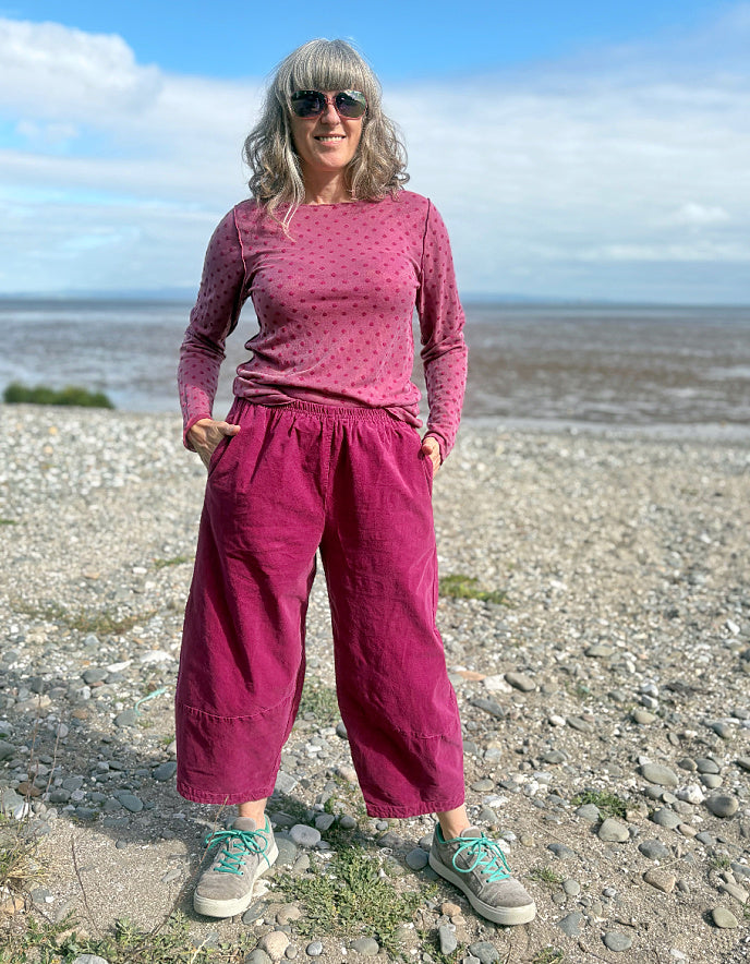 Woman in pink needlecord lantern trousers and a pink pink spotty top standing on a pebbly beach with ocean and sky in the background