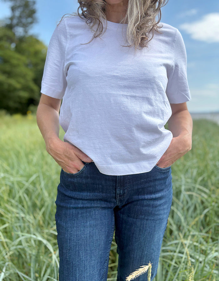 Person wearing a white t-shirt and blue jeans standing in a grassy field with a clear sky.