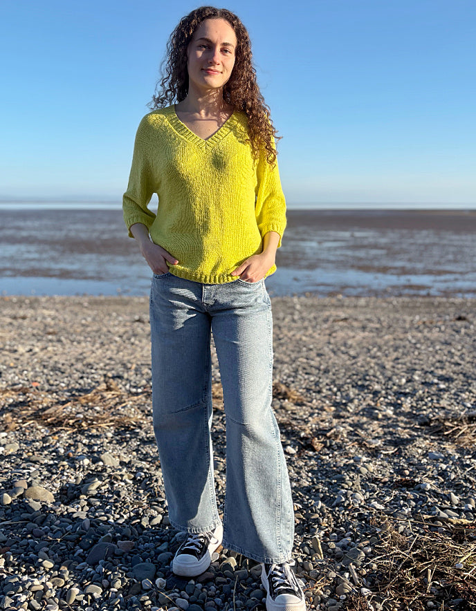 Woman in a yellow sweater and blue jeans standing on a pebbly beach with water and sky in the background.