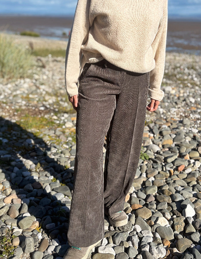 A woman standing on a pebble beach wearing dark brown wide-leg corduroy pants, a cream knit sweater, and dark sneakers, with water and sky in the background.