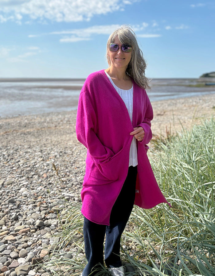 Person wearing a bright pink long wool cardigan standing on a pebbly beach with grass and water in the background.