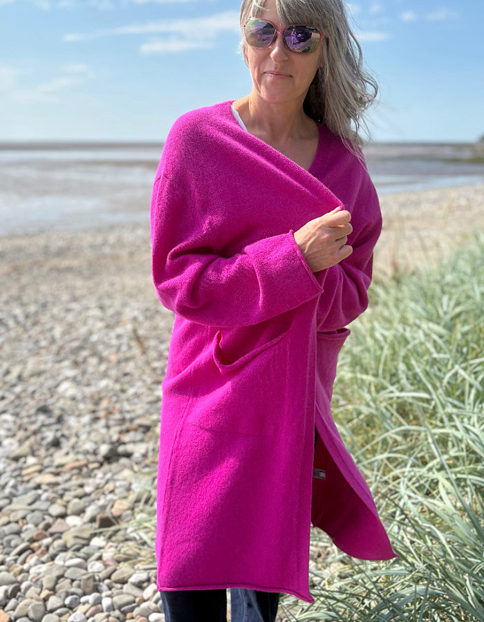 Person wearing a bright pink long wool cardigan standing on a pebbly beach with grass and water in the background.