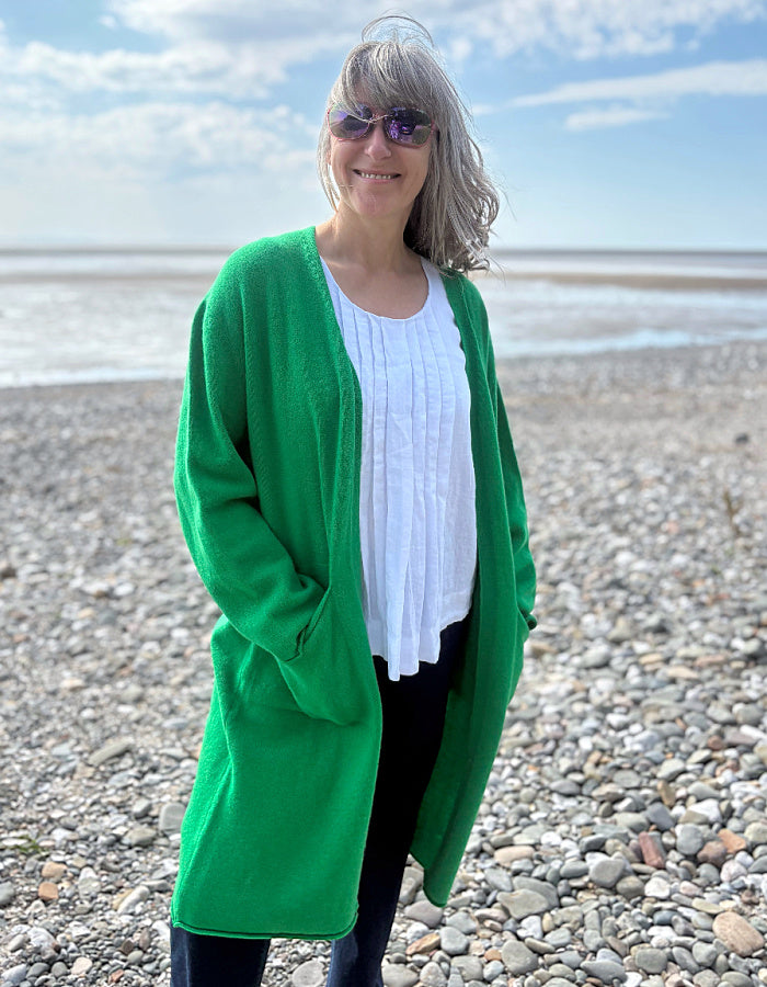 Person wearing a green wrap long merino wool cardigan on a pebbly beach with ocean and sky in the background