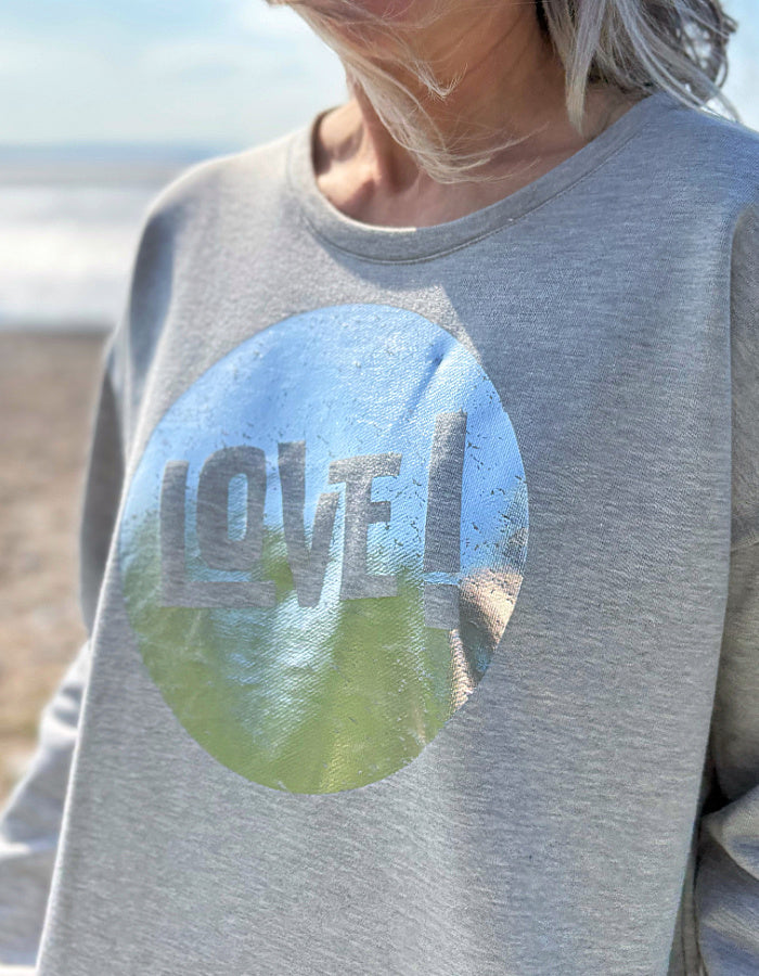 Gray sweatshirt with a circular design featuring 'LOVE' and nature imagery, worn by a person outdoors.