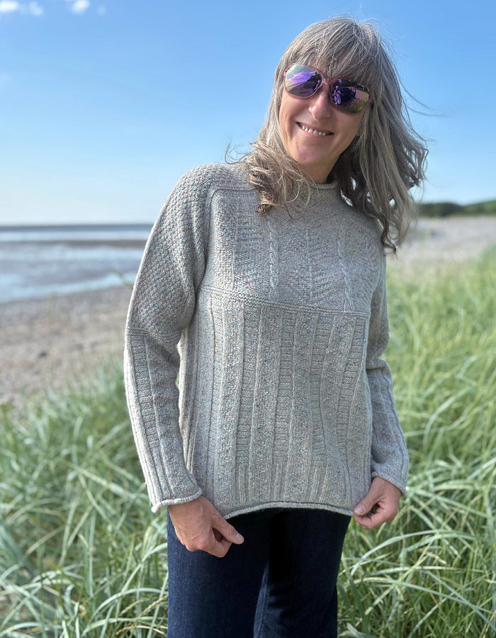 Person wearing a donegal tweed wool grey sweater with wide leg jeans and sunglasses on a beach