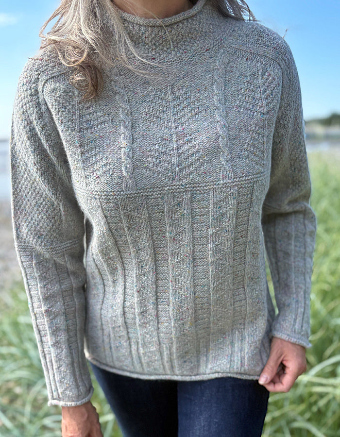 Person wearing a donegal tweed wool grey sweater with wide leg jeans and sunglasses on a beach