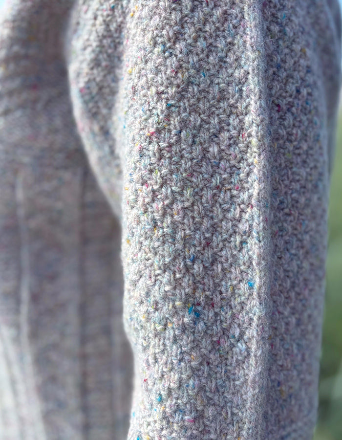 Person wearing a donegal tweed wool grey sweater with wide leg jeans and sunglasses on a beach