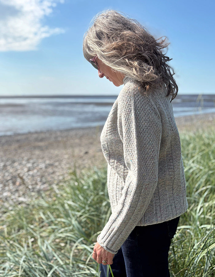 Person wearing a donegal tweed wool grey sweater with wide leg jeans and sunglasses on a beach