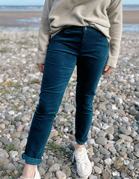 A woman standing on a rocky beach wearing a light beige knit sweater, dark teal velvet pants, and white sneakers, with the ocean and blue sky in the background.