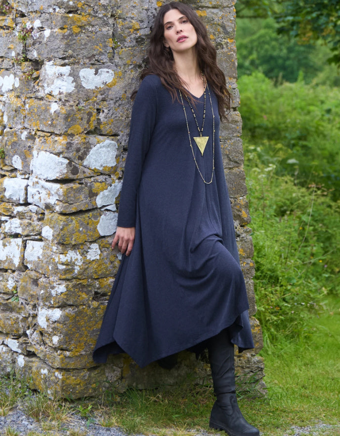 Woman in a long dark blue dress standing against a stone wall with greenery in the background