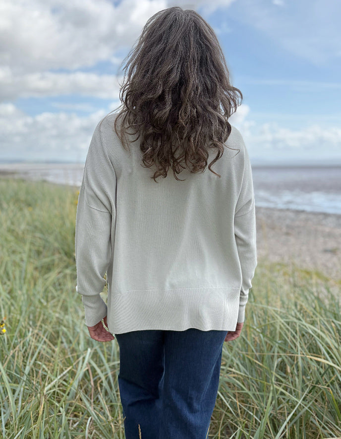 Woman standing in beach grass wearing a light-coloured cream turtle neck sweater and sunglasses with a beach and sky in the background.
