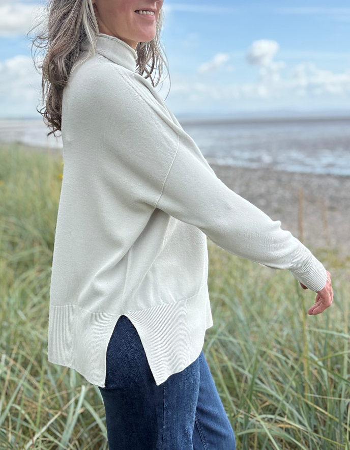 Woman standing in beach grass wearing a light-coloured cream turtle neck sweater and sunglasses with a beach and sky in the background.