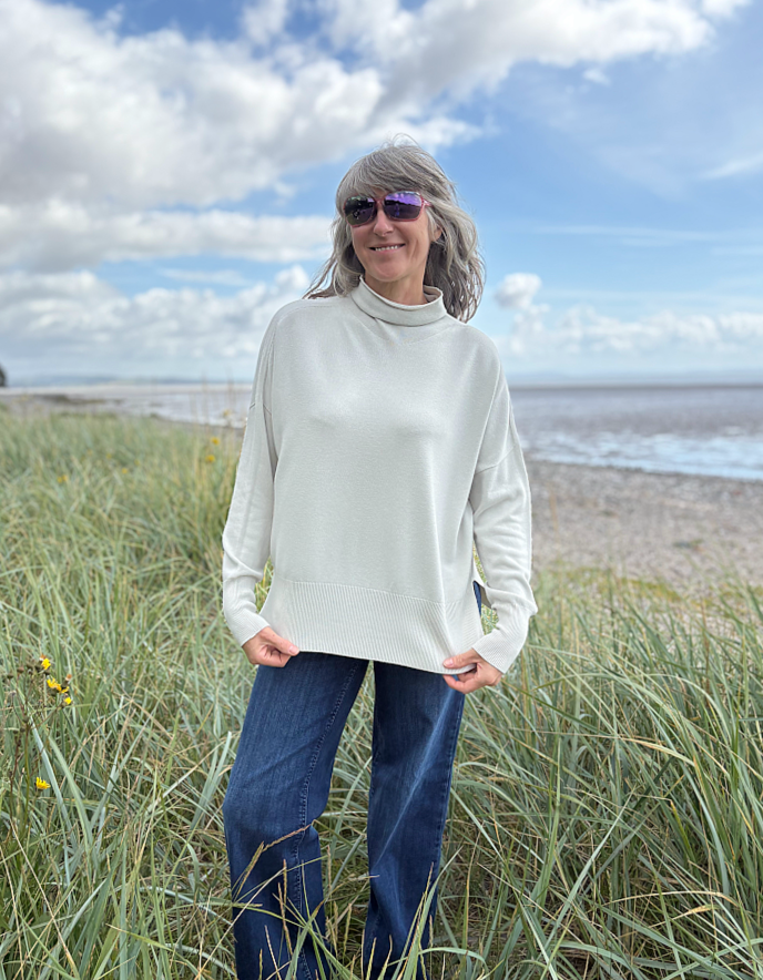 Woman standing in beach grass wearing a light-coloured cream turtle neck sweater and sunglasses with a beach and sky in the background.