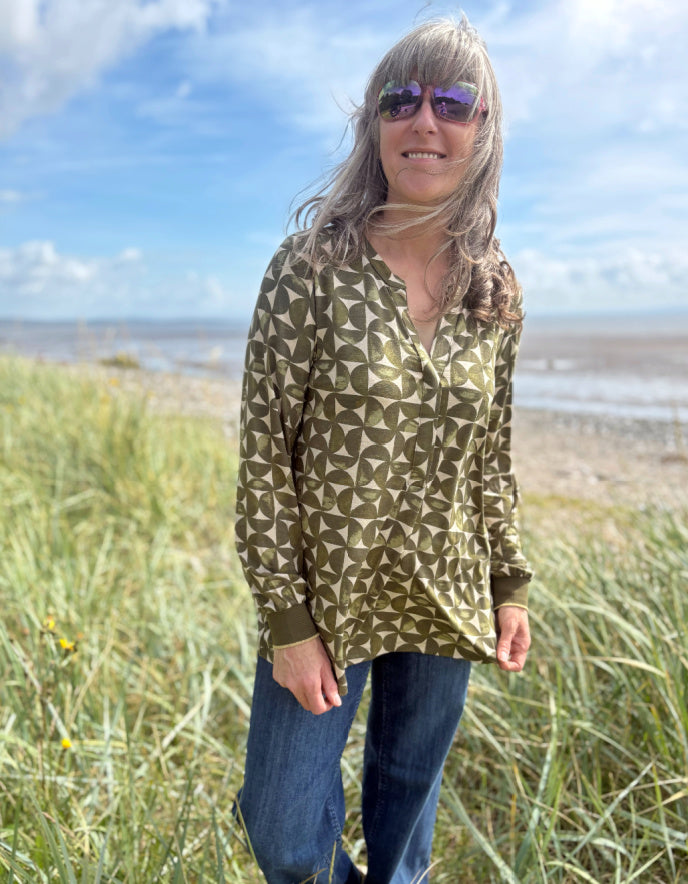 Woman wearing a olive geopatterned shirt and sunglasses standing in grass with a beach in the background