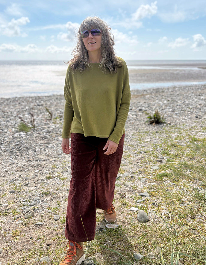 Person standing on a beach wearing a green sweater and brown pants.