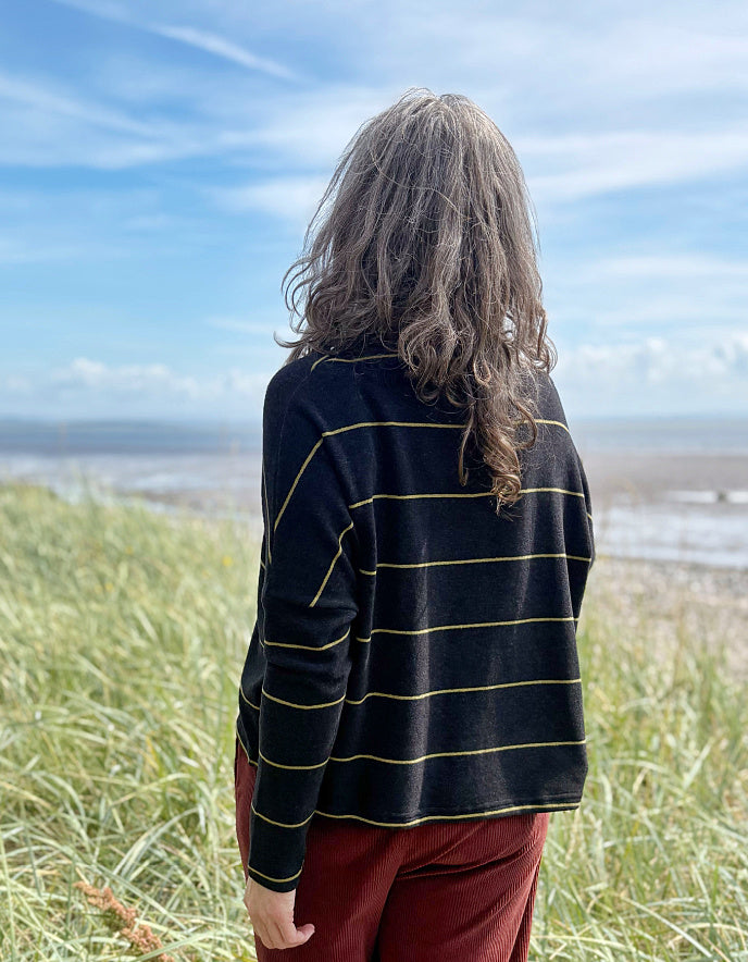 Person wearing a black and yellow striped shirt standing on grassy dunes with a beach and blue sky in the background