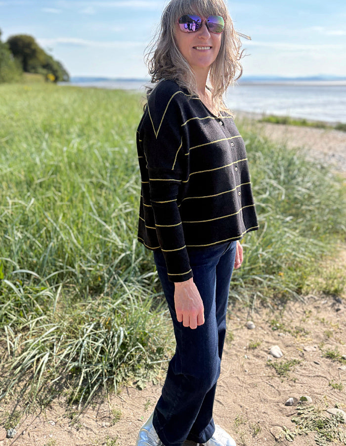 Woman wearing charcoal and olive pin stripe cardigan and sunglasses, standing in a grassy field with a body of water in the background.