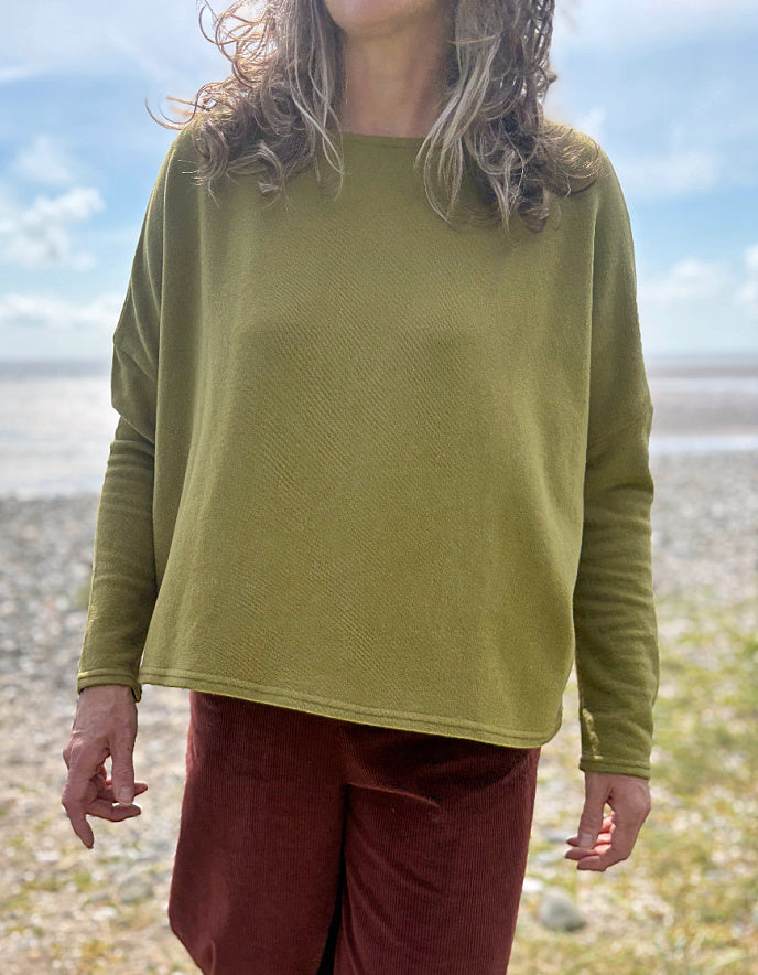 Person wearing a green textured sweater and red pants standing on a beach.