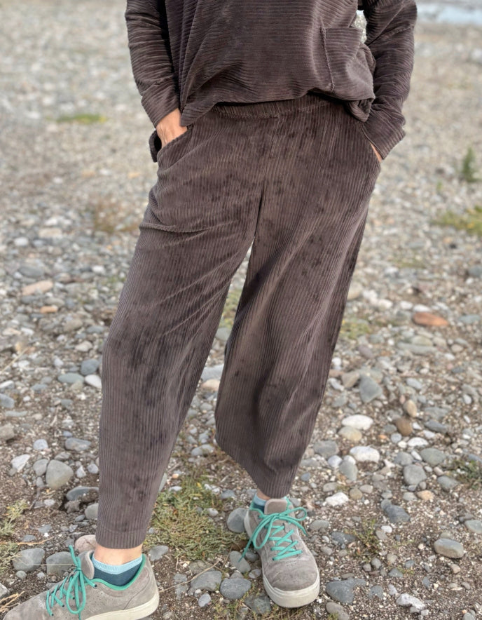 Person wearing a brown cord lantern shaped trousers standing on a pebbly beach with ocean in the background