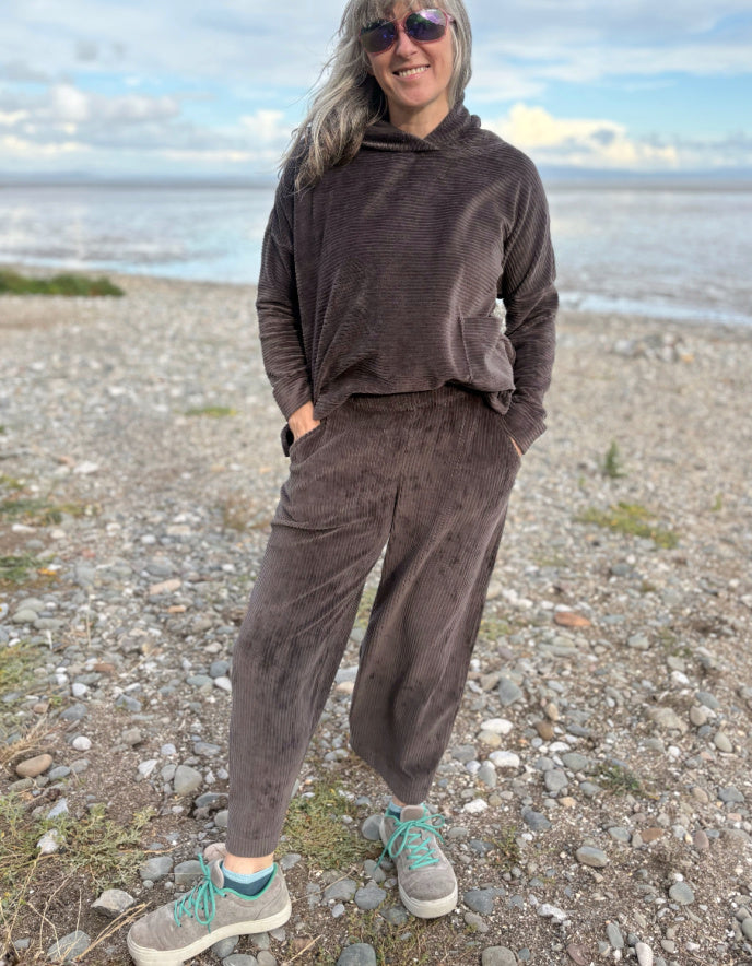 Person wearing a brown cord lantern shaped trousers standing on a pebbly beach with ocean in the background