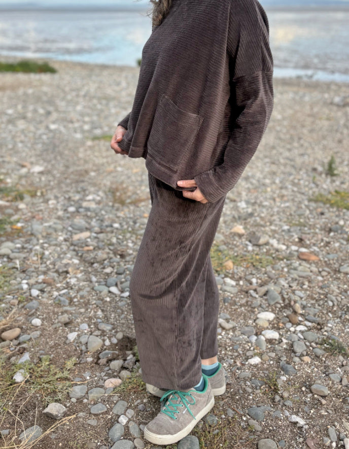 Person wearing a brown cord lantern shaped trousers standing on a pebbly beach with ocean in the background