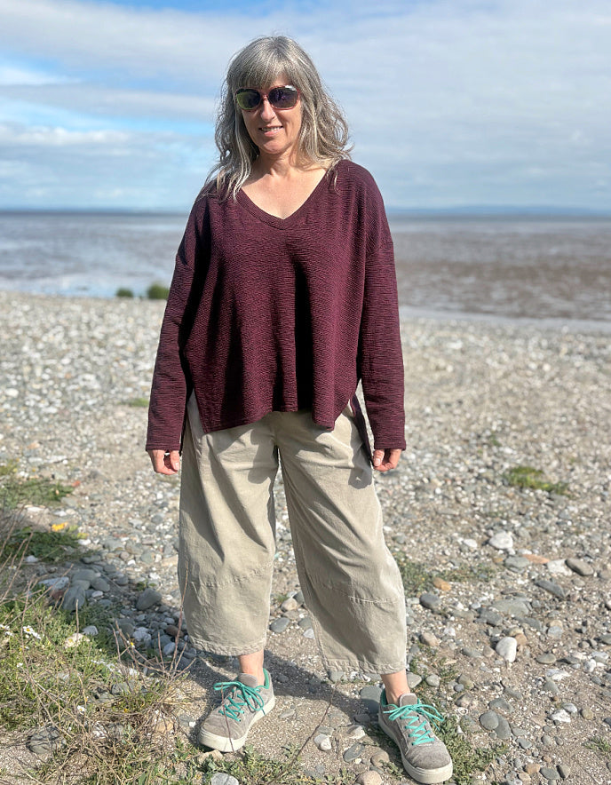 Woman standing on a pebbly beach wearing a burgundy sweater and beige pants.