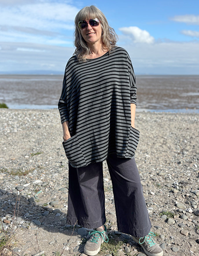 Woman standing on a pebbly beach wearing a striped black and grey oversized sweatshirt top and sunglasses.