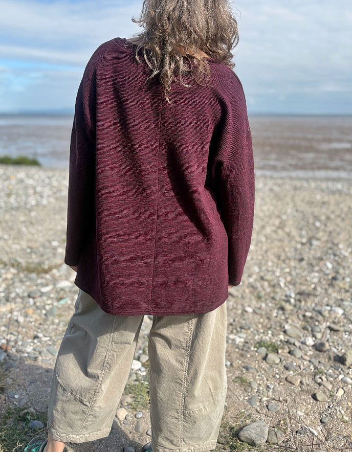 Person wearing a maroon sweater and beige pants standing on a pebbly beach.