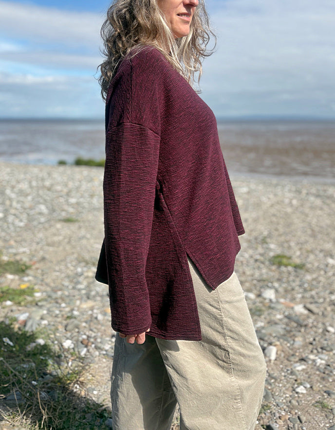 Person wearing a maroon sweater and beige pants standing on a pebbly beach with water and sky in the background.