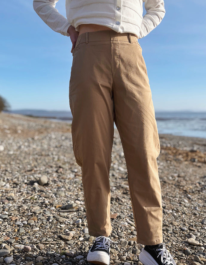 Person wearing beige pants and a white jacket standing on a pebbly beach with ocean view.