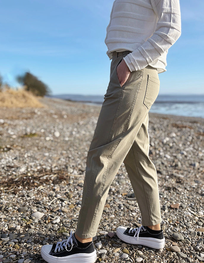 Person wearing beige pants and black sneakers on a pebbly beach with a blue sky.