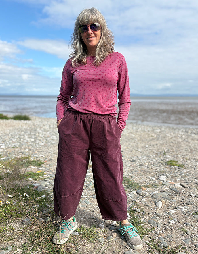 Woman in pink top and maroon pants standing on a pebbly beach with ocean and sky in the background