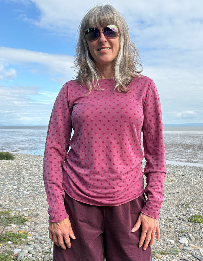 Woman wearing a pink long-sleeve top and purple pants standing on a beach with blue sky and water in the background.