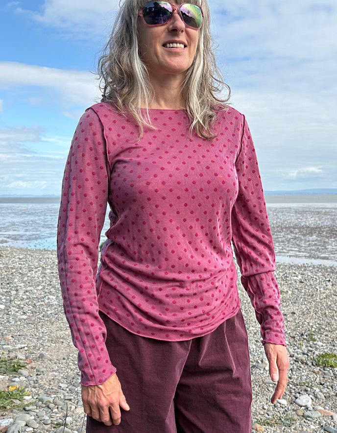 Woman wearing a pink long-sleeve top with a pattern and purple pants standing on a beach.