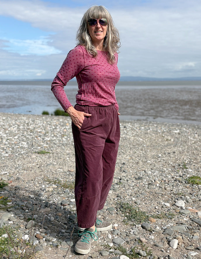 Woman standing on a pebbly beach wearing a pink top and purple pants.