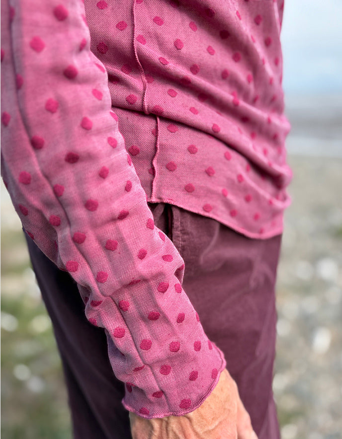 Person wearing a pink long-sleeve shirt with red polka dots and purple pants on a blurred natural background.