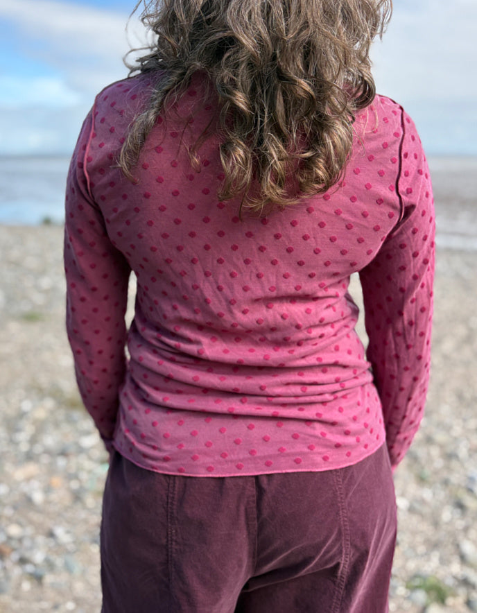 Person wearing a pink polka dot shirt and purple pants standing on a beach.