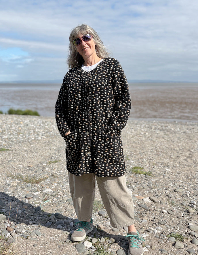 Person standing on a pebbly beach wearing a black felted wool patterned coat.