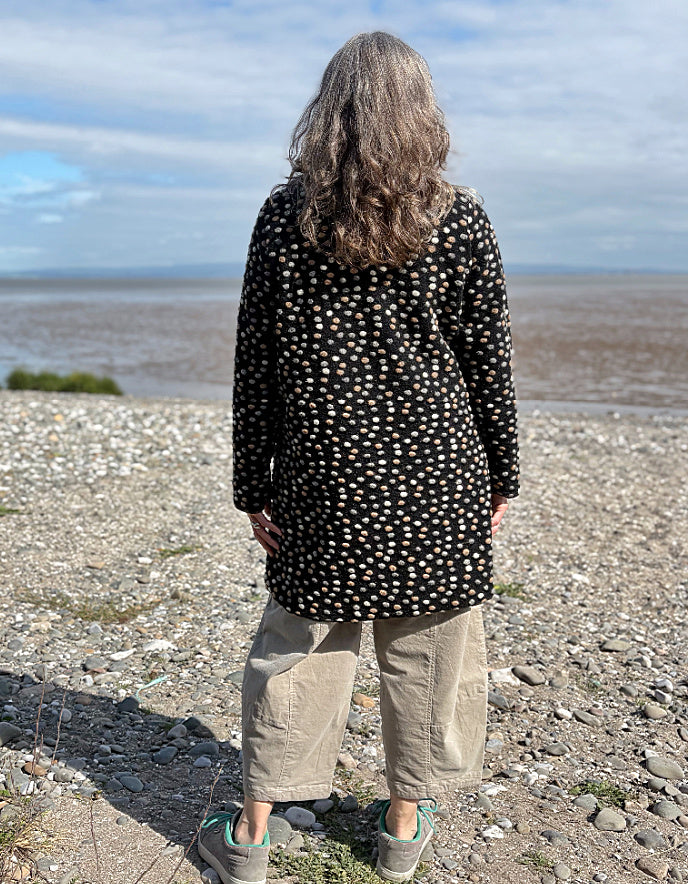 Person wearing a black polka dot coat standing on a pebbly beach with a vast landscape in the background.