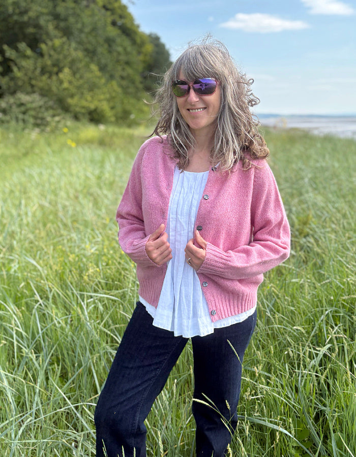 Woman wearing a short pink wool cardigan in a grassy field with a scenic background.
