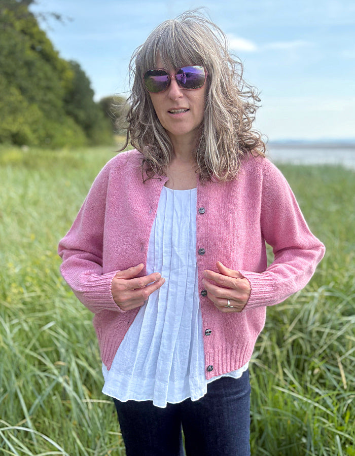 Woman wearing a short pink wool cardigan in a grassy field with a scenic background.