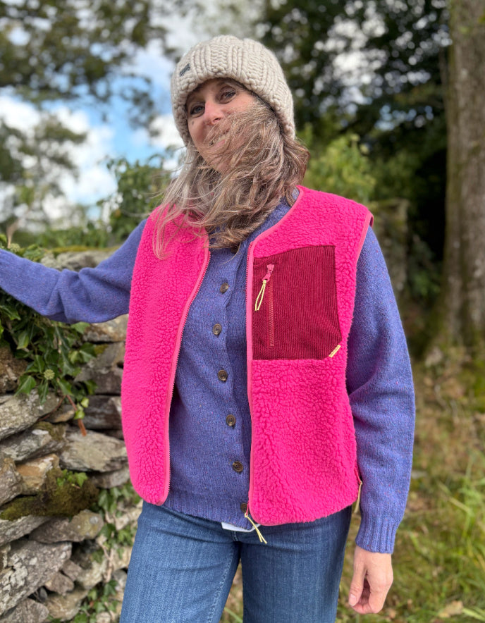 Person wearing a pink vest over a blue sweater outdoors near a stone wall.