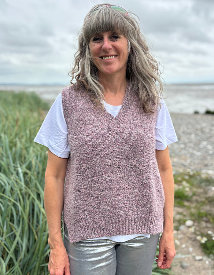 Woman wearing a pink knitted vest over a white shirt with a beach and grass background
