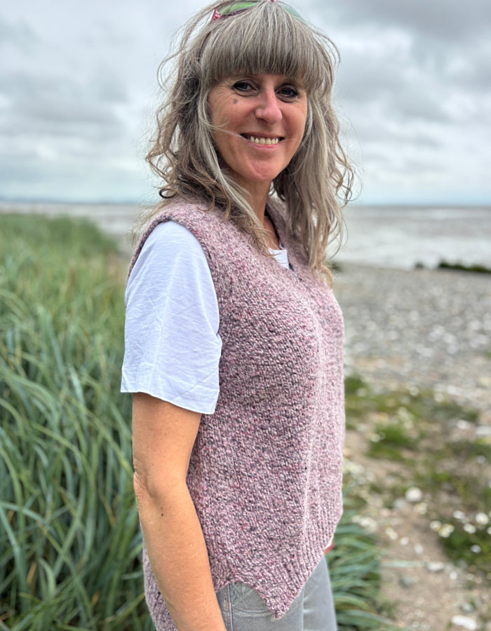 Woman wearing a pink knitted vest standing on a beach with grass and water in the background