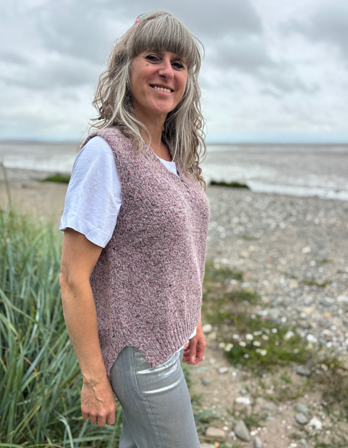 Woman standing on a beach wearing a pink knitted vest and white shirt.