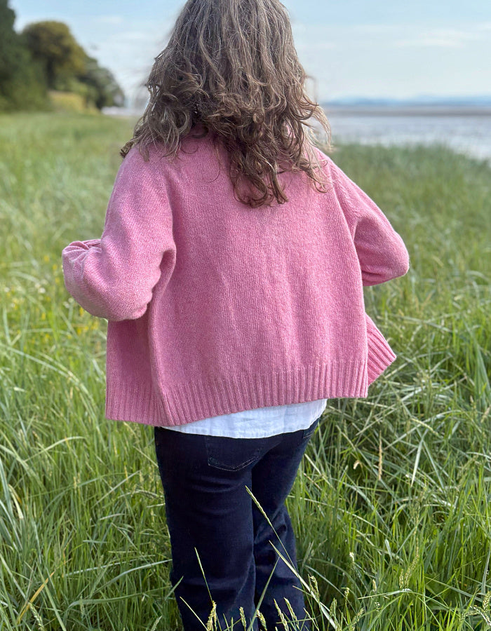 Woman wearing a short pink wool cardigan in a grassy field with a scenic background.