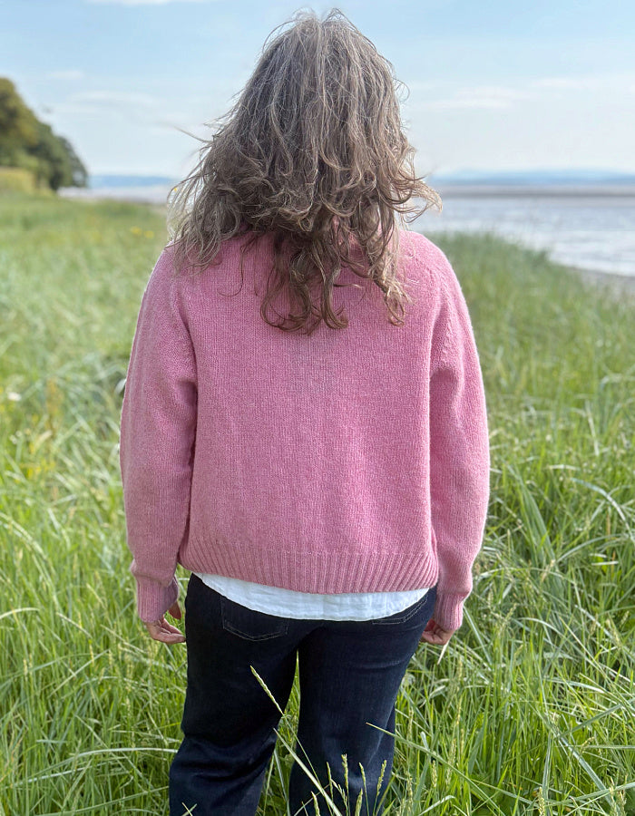 Woman wearing a short pink wool cardigan in a grassy field with a scenic background.