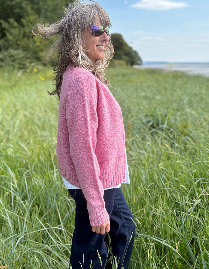 Woman wearing a short pink wool cardigan in a grassy field with a scenic background.