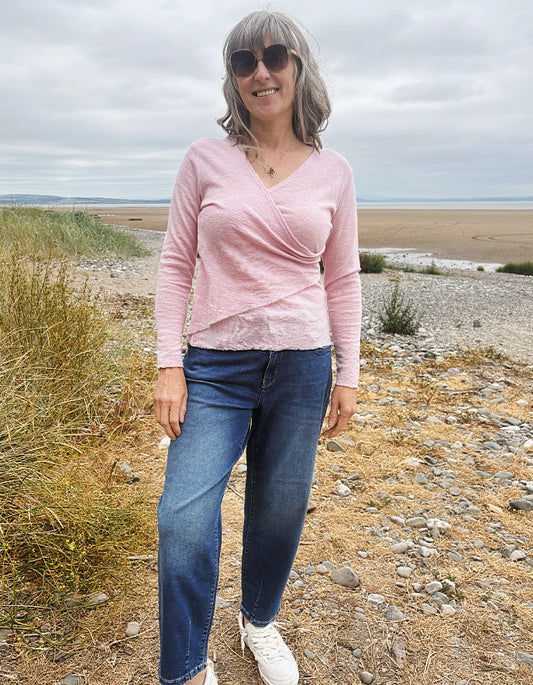 A woman standing outdoors on a rocky beach path wearing a light pink long-sleeve wrap-style top, blue jeans, and white sneakers.